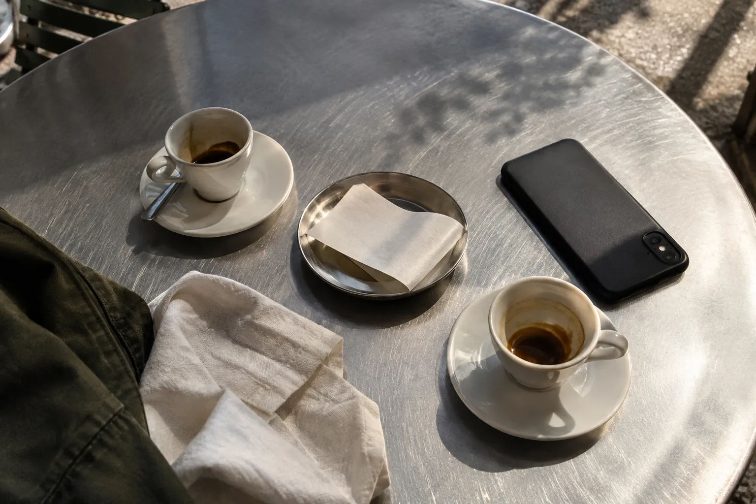 Two coffees, a folded receipt and a phone on a sunlit café table.